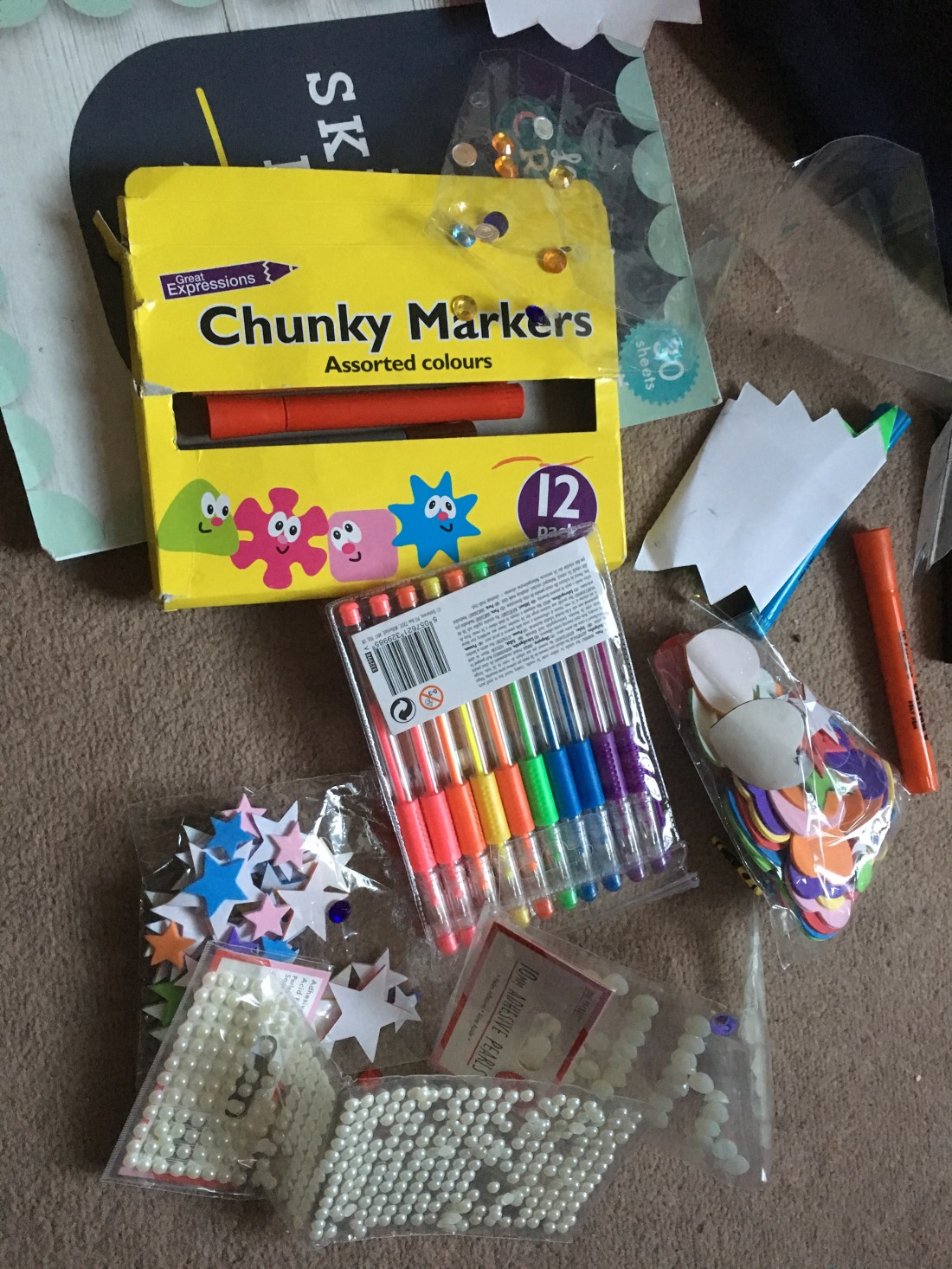 A photograph of a box of chunky crayons, some colouring pens, and a small assortment of craft stickers all on a table together.